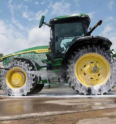 A person in waterproof gear uses a pressure washer to clean mud from the large tire and hood of a green tractor.