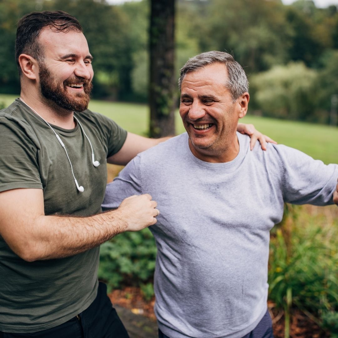 Two men, smiling, outdoors; one assists the other with arm stretches.