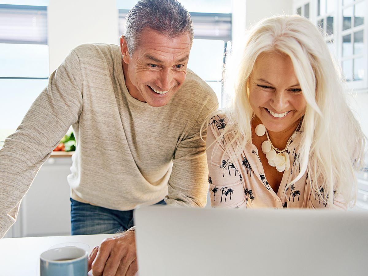 Couple smiling at laptop in a kitchen, man leaning over, woman seated.
