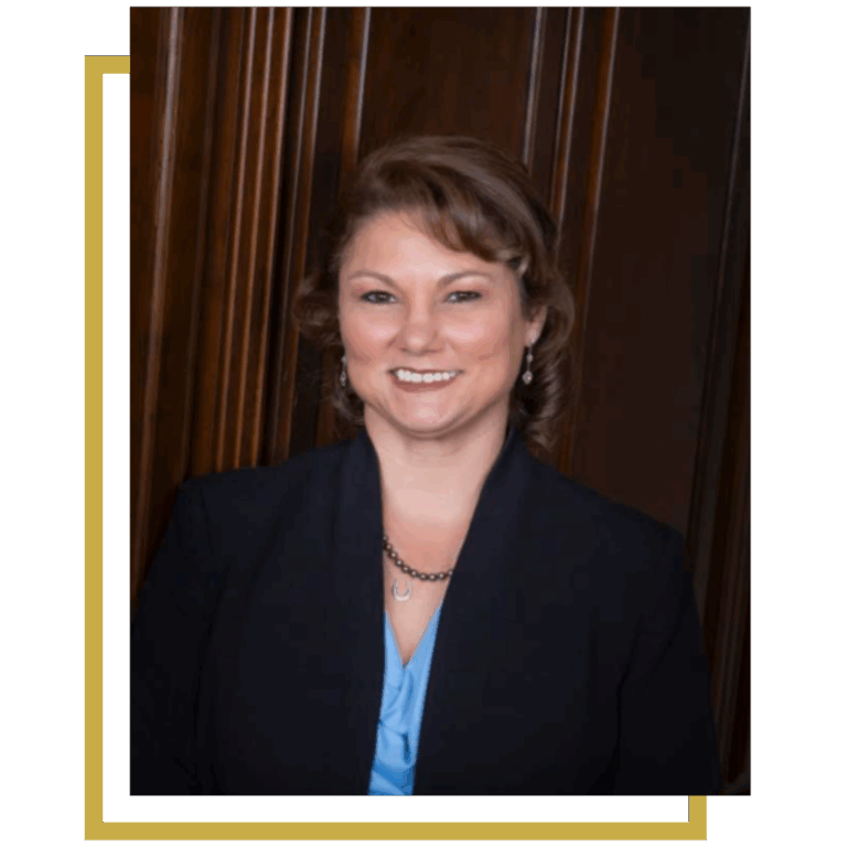 Woman in a blue blouse and navy blazer smiles, standing in front of wood paneling.