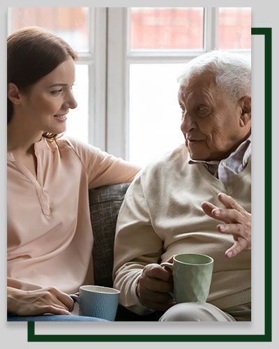 Woman and older adult sitting on a couch, talking. Holding mugs, in front of a window.
