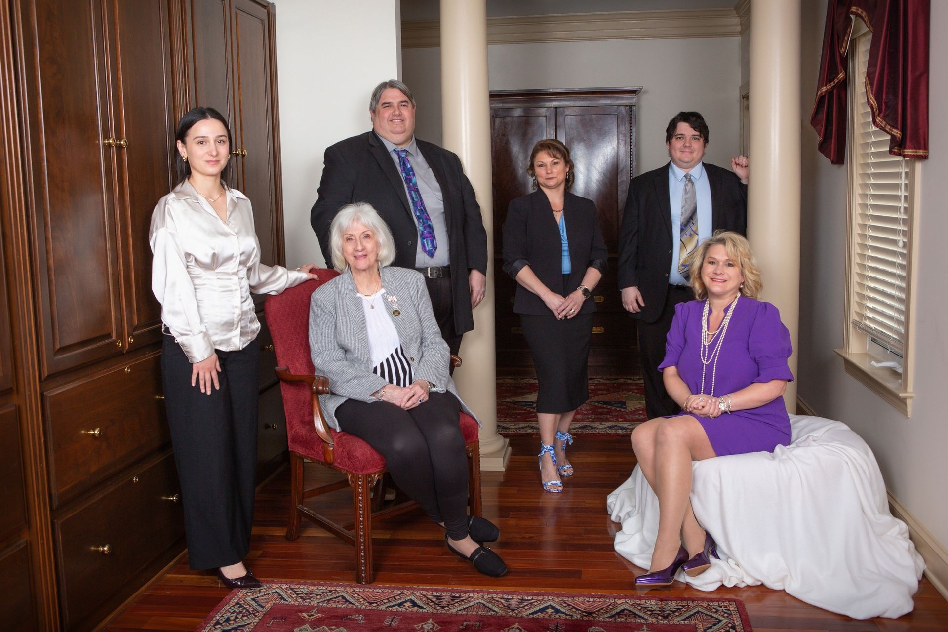 Group of people posing in an interior. Men and women wear business attire. Setting has pillars, wood cabinetry and rug.