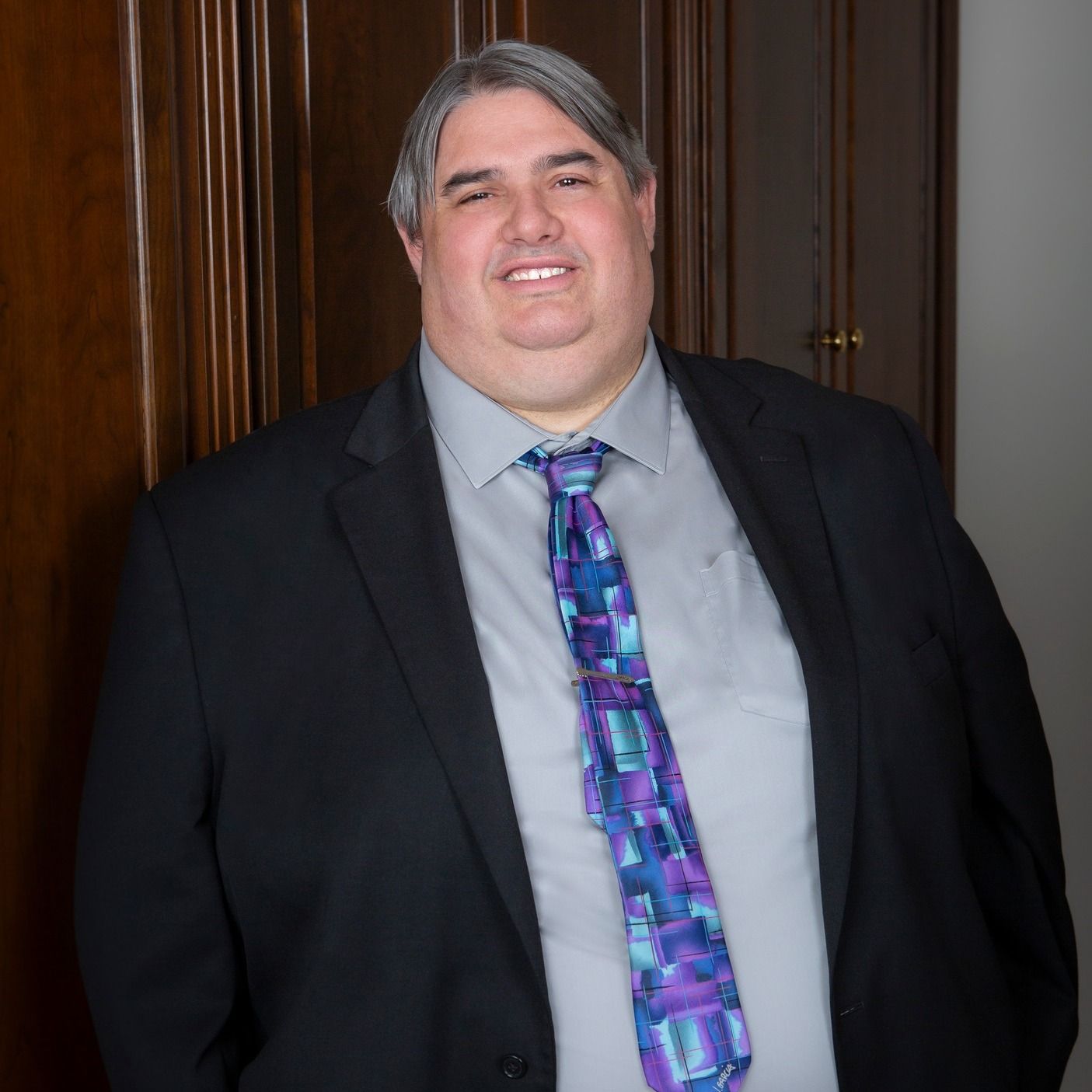 Man in a suit smiles, standing near a wood-paneled wall, wearing a patterned tie and gray shirt.