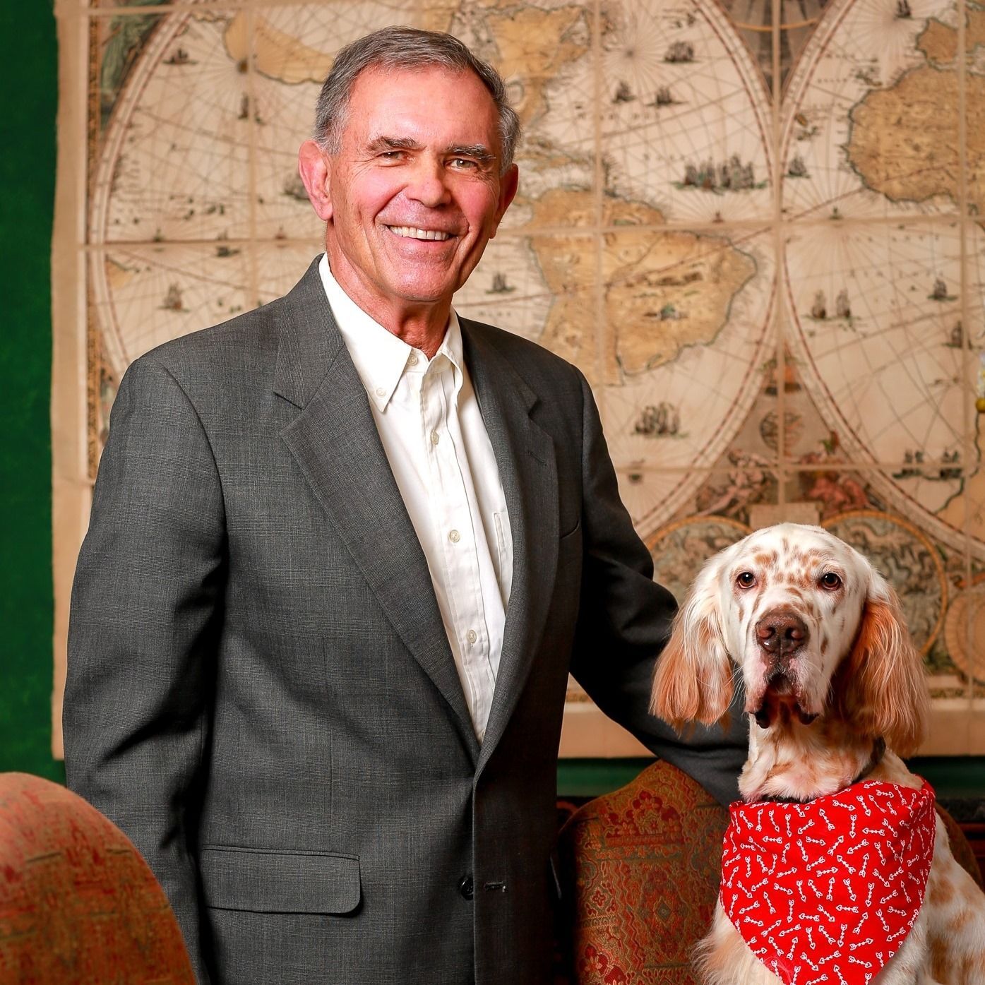 Man in suit smiles, standing with a dog wearing a bandana, in front of a world map.