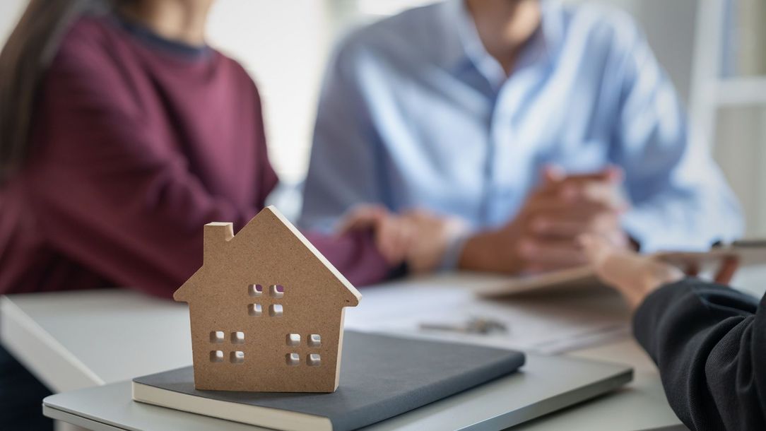 Wooden house model sits on a notebook in front of a couple at a table.
