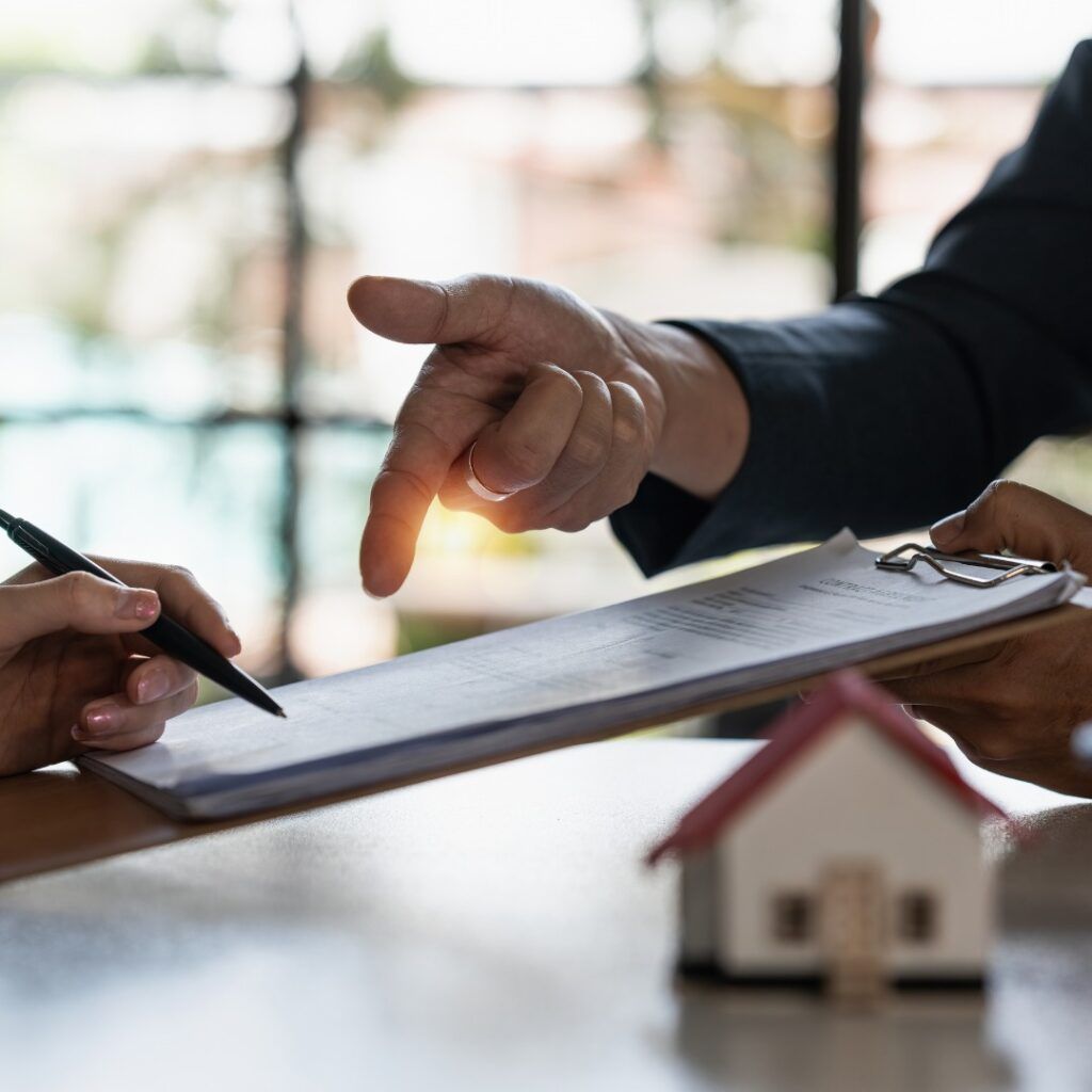 Person signing a document, being pointed to by another person, model house on table.