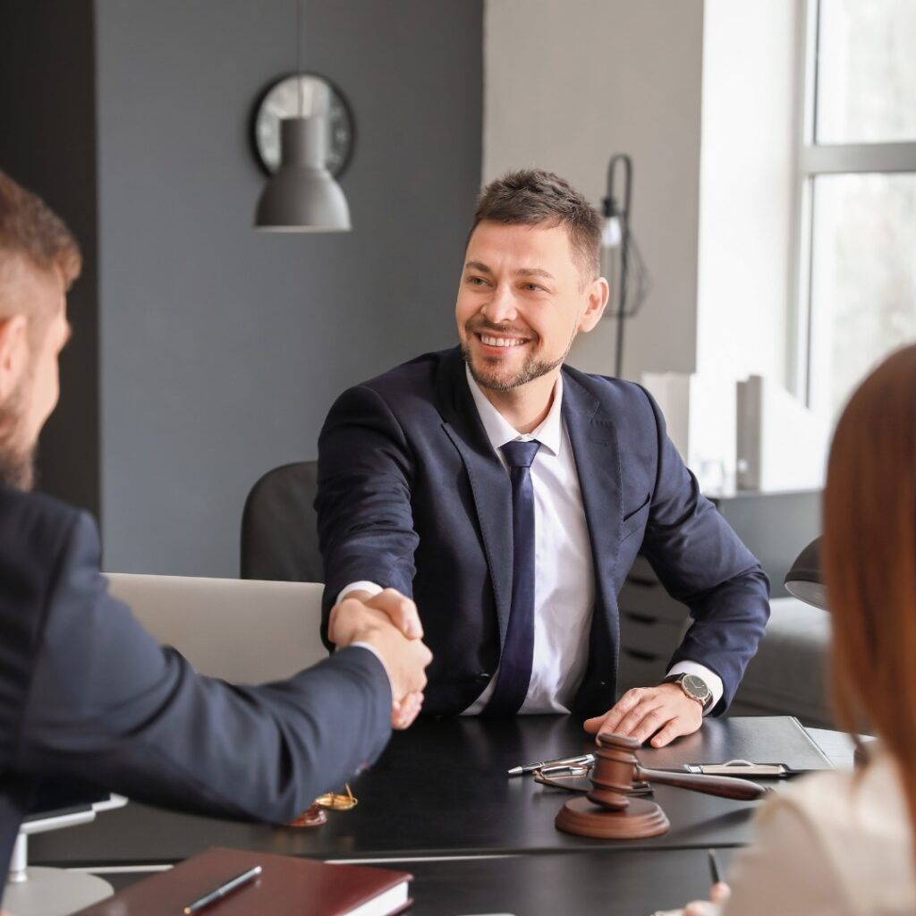 Man in suit shakes hands with another man at a desk; a woman and gavel are also present.