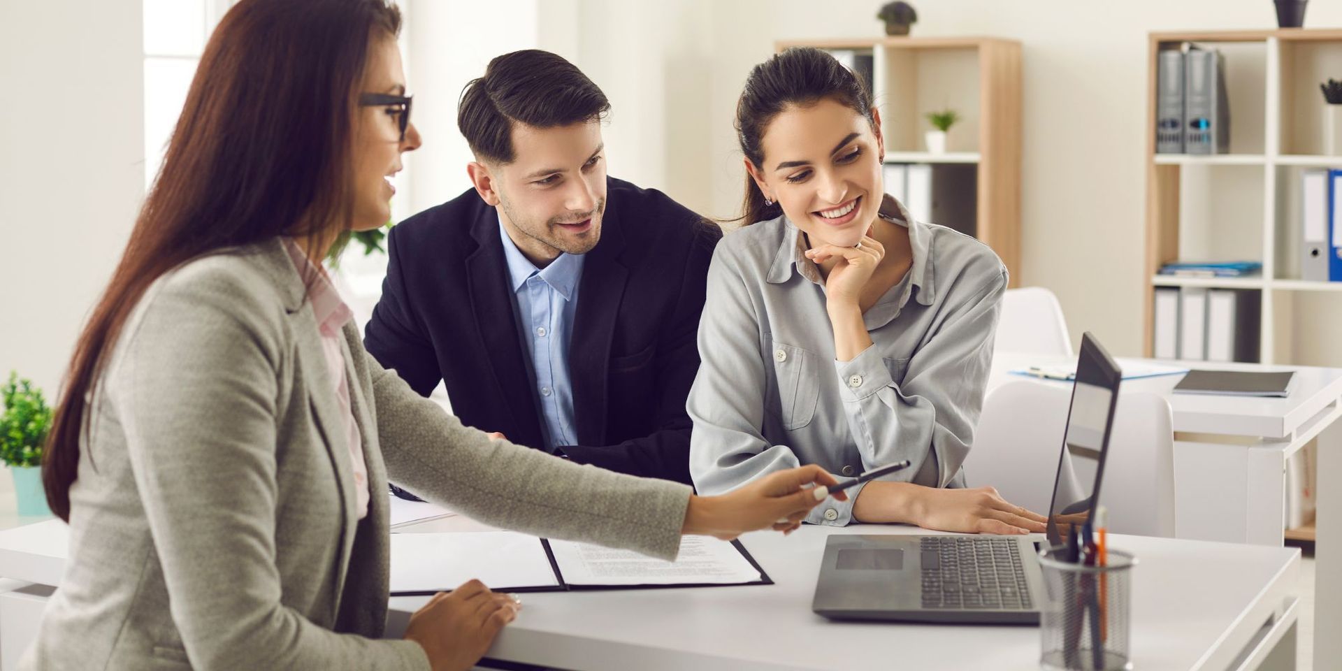 Three people reviewing a laptop at a desk in an office setting.