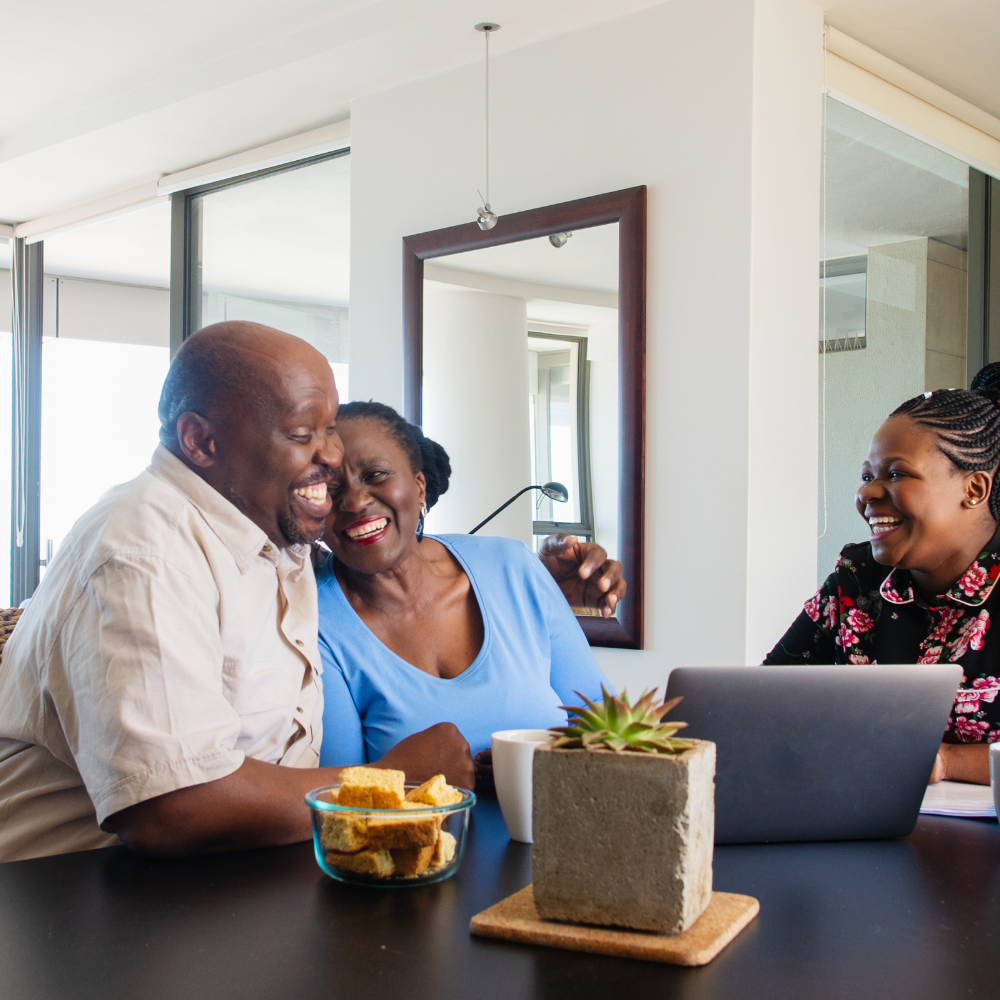 Smiling couple embraces, looking at laptop with another woman. Table with snacks, plant. Bright room.