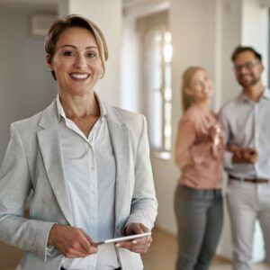 Real estate agent smiling, holding tablet, with potential buyers in the background.