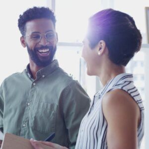 Two people laughing, looking at each other. Man with glasses, holding a notepad. Woman in striped shirt. Bright setting.
