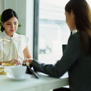 Two women at a table, one in white and one in black, possibly discussing legal documents.
