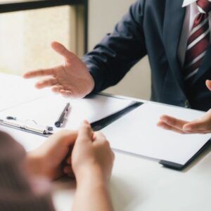 Two people at a desk; one in a suit gestures, the other reviews a document.