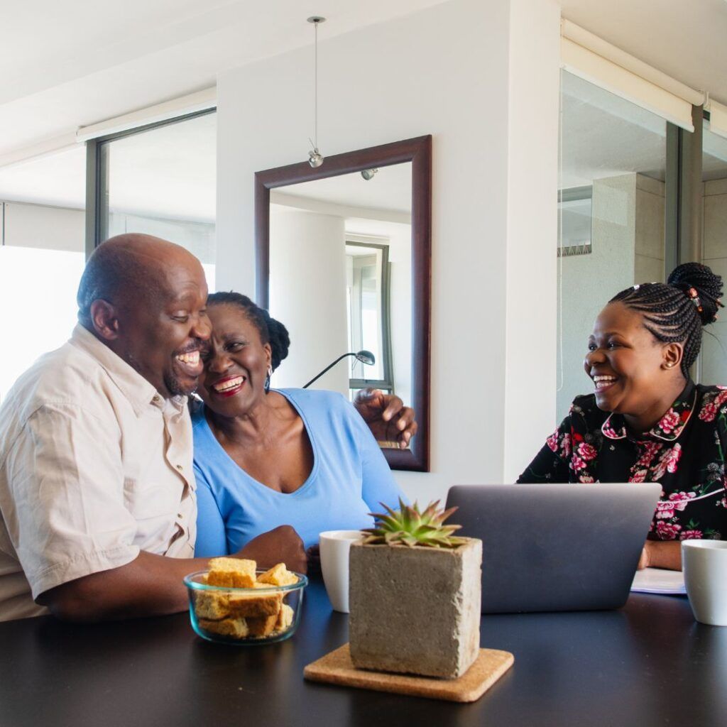 Senior couple laughs with a woman at a table with laptop, snacks, and drinks.