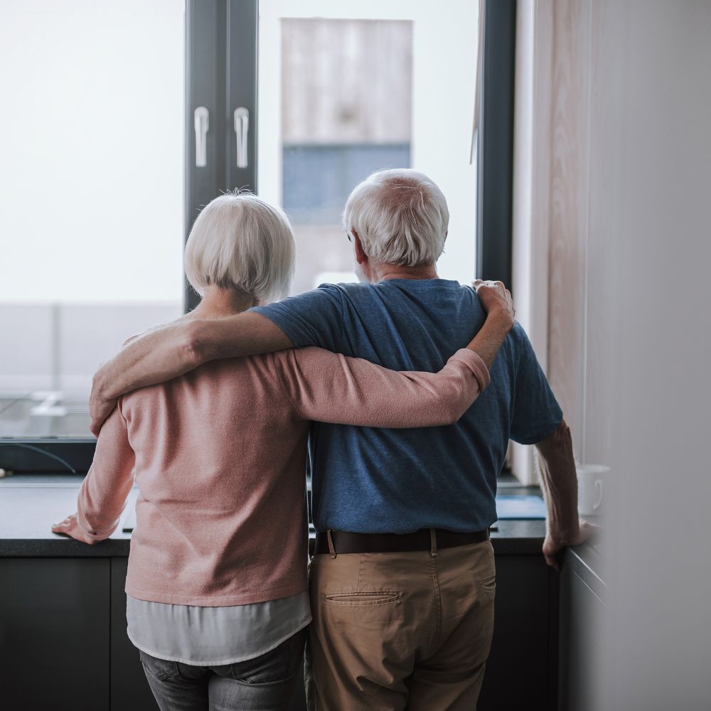 Couple, arms around each other, looking out a window. They are both wearing casual clothes, standing indoors.