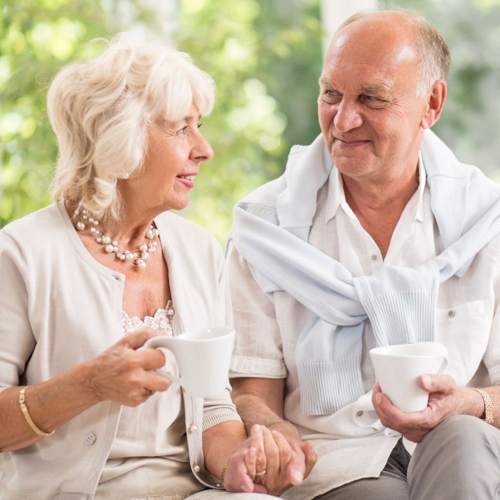 Elderly couple holding hands, sipping from white mugs. Woman looks at man, smiling; man looks back, smiling. Outdoors.