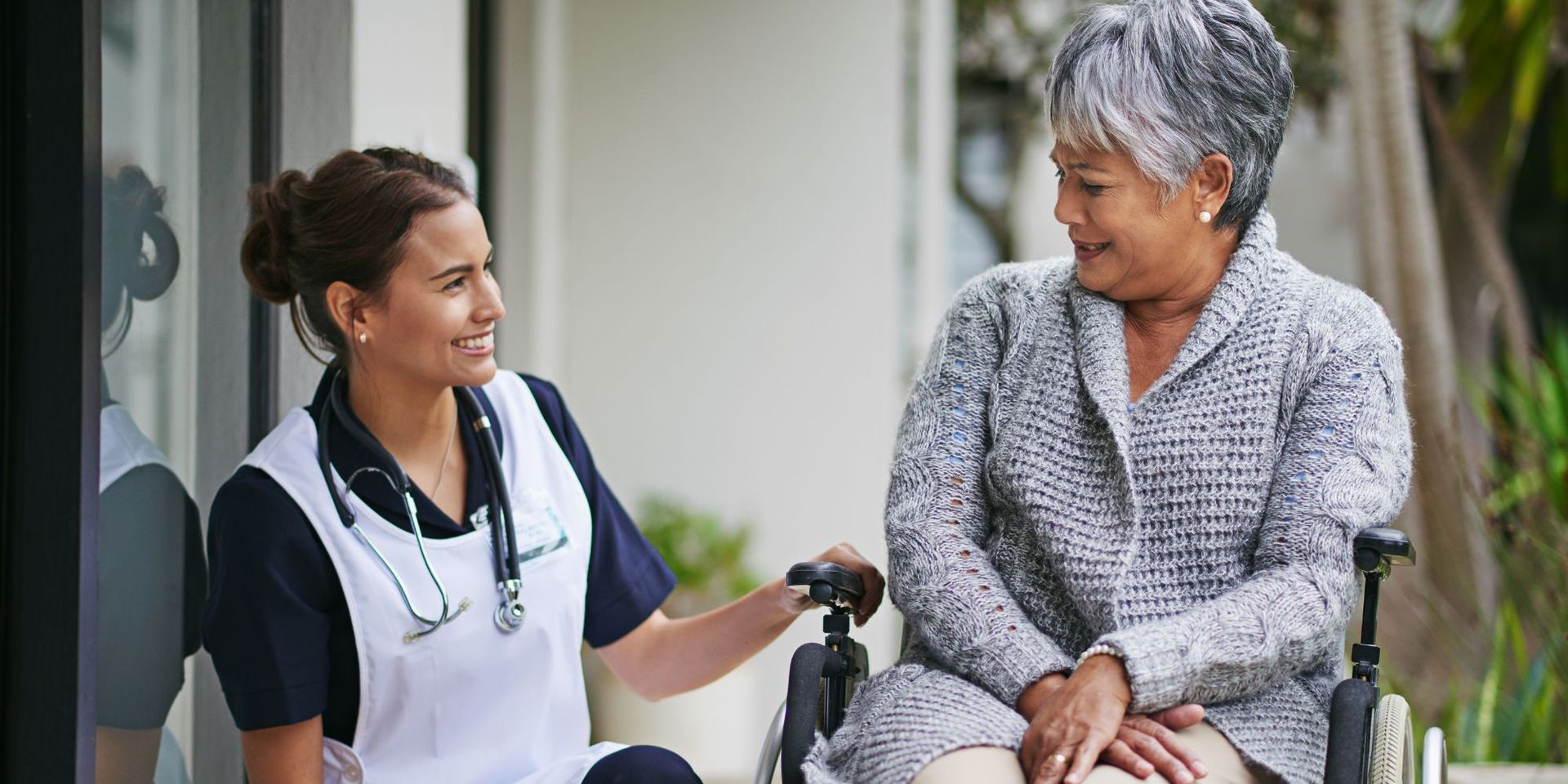A caregiver in a white apron with a stethoscope and an older person in a wheelchair are smiling and talking outdoors.