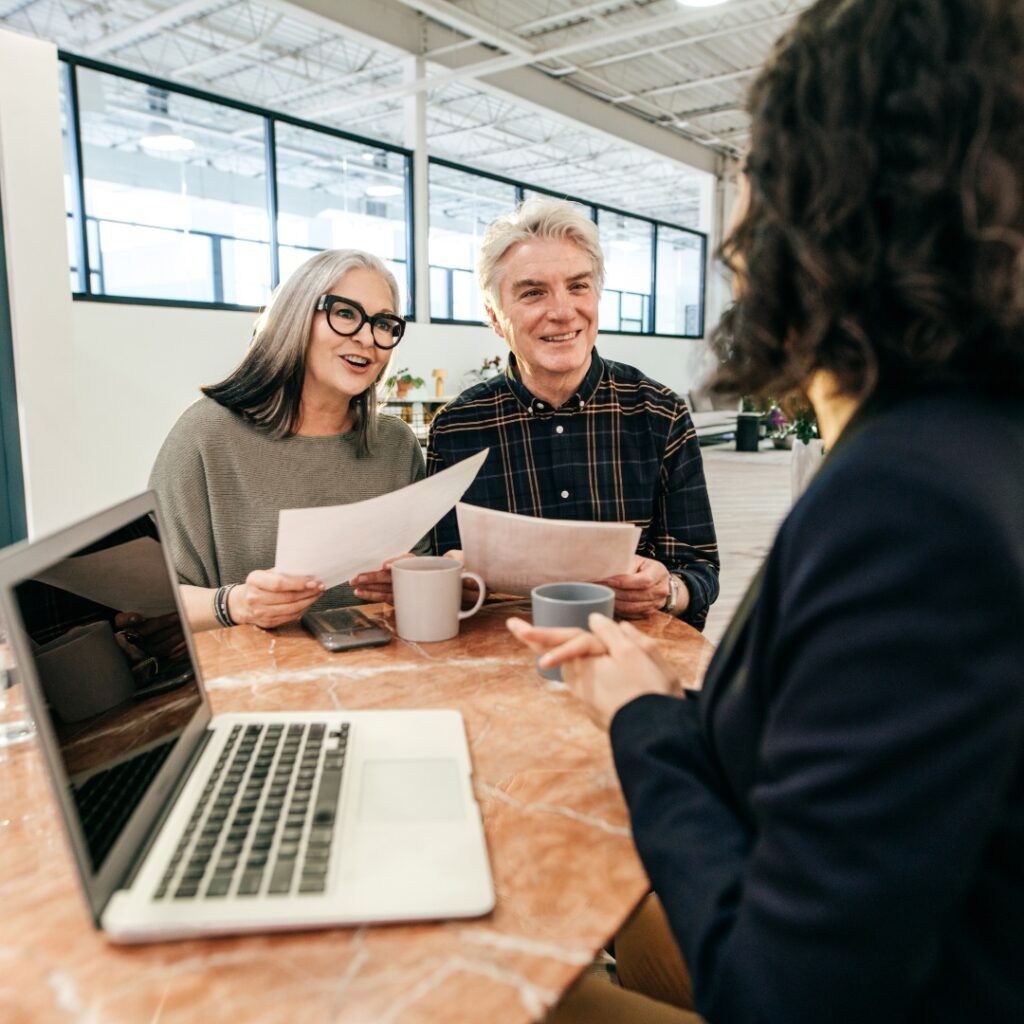 A couple consults with a person at a table, reviewing documents and using a laptop in a modern office.