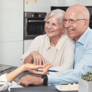 Elderly couple smiling as someone points to a document; kitchen setting.