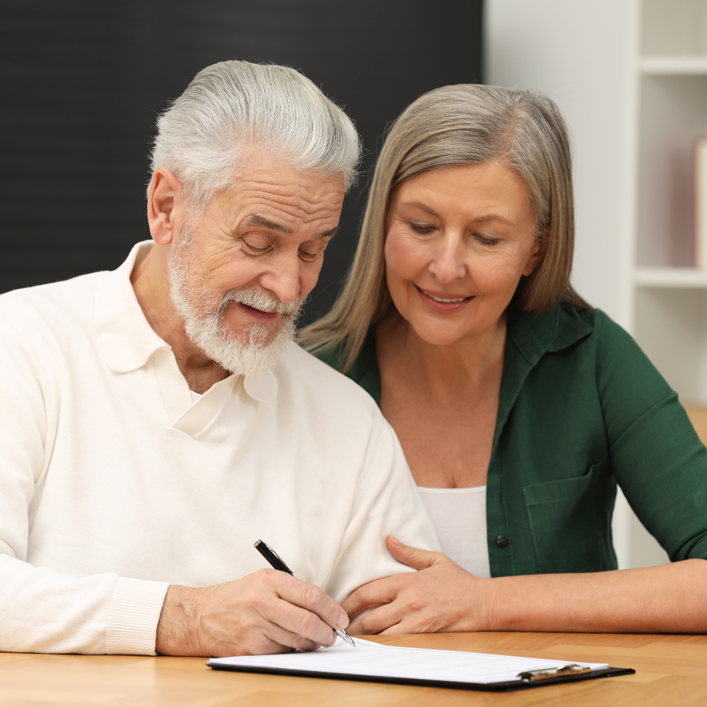 Older person signing document, assisted by another; indoor setting.