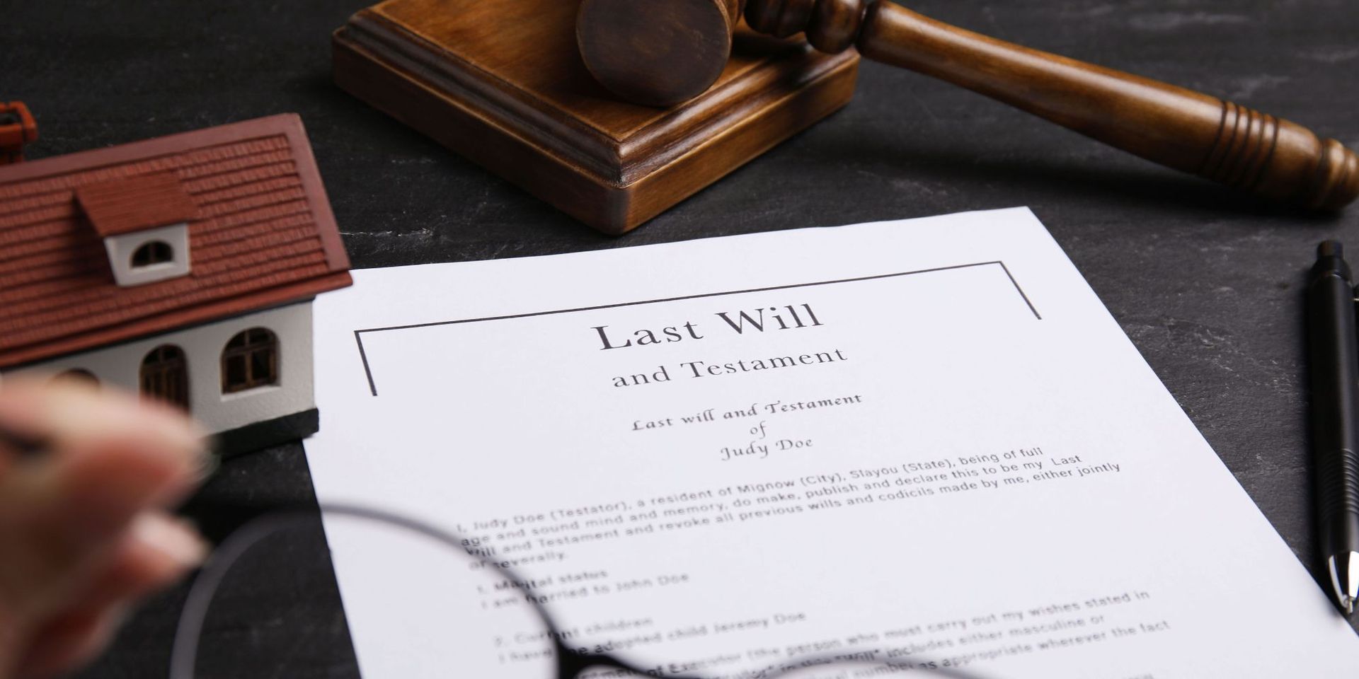 A Last Will and Testament document, a toy house, a gavel, and a pen on a dark surface.
