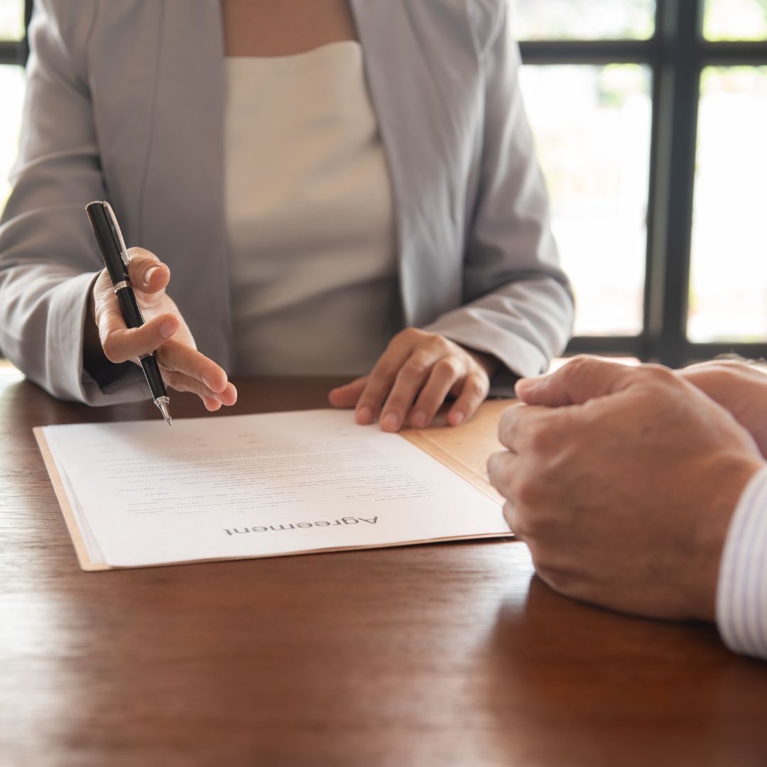 Woman in blazer pointing to document, discussing with person at wooden table.