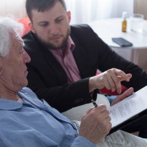 Man in a suit pointing at document held by elderly man in bed, indoors.
