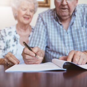 Elderly couple signing document at a table, man holding pen, woman smiling.