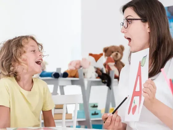 Therapist with glasses holds letter card, child imitates sound, in a room with toys and art supplies.