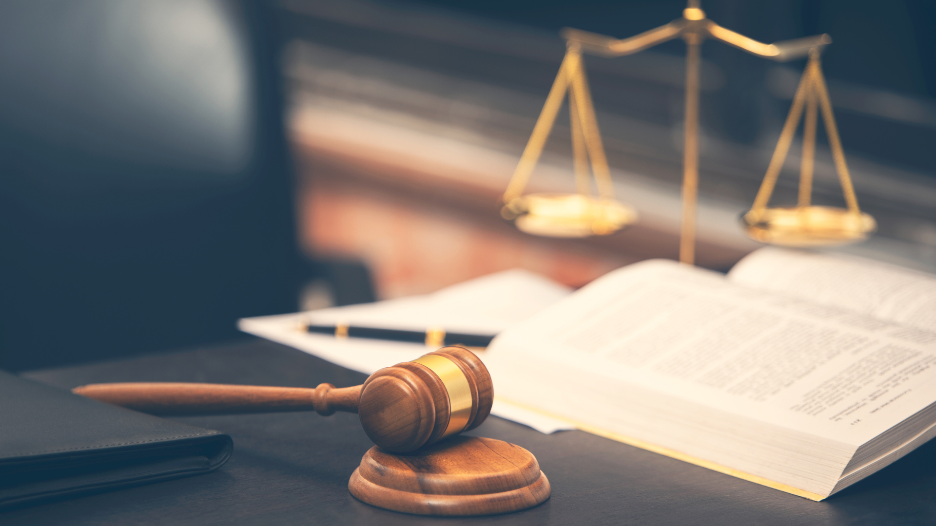 Gavel on desk in front of open book and scales, symbols of law and justice.