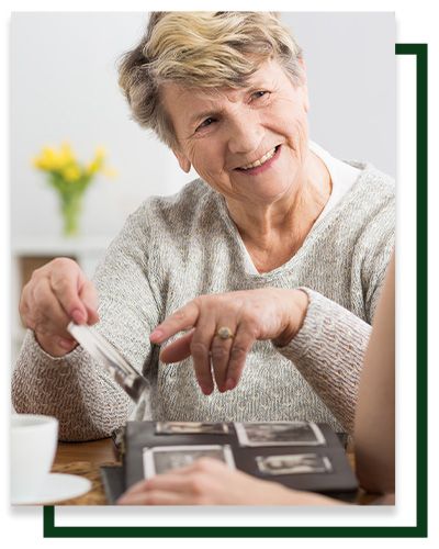 Woman looking at photo album, smiling. Holding a card, seated indoors, near a cup and flowers.
