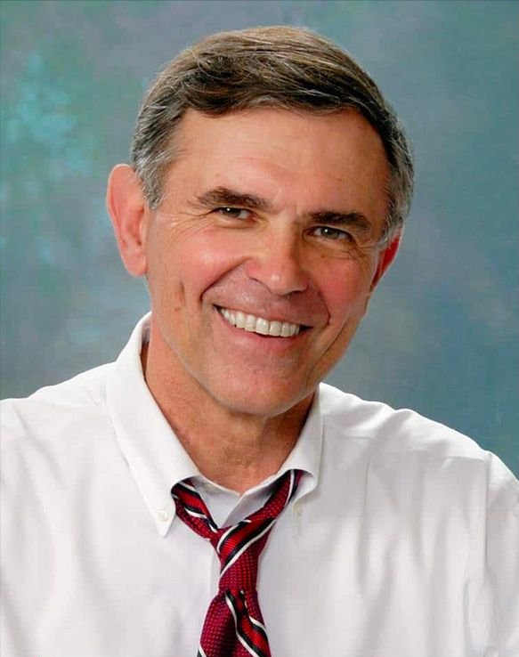 Man in white shirt with red striped tie smiles, against a blurred background.
