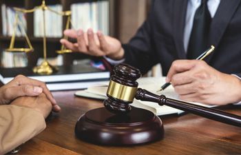 Wooden gavel on table; legal consultation with scales of justice in background.