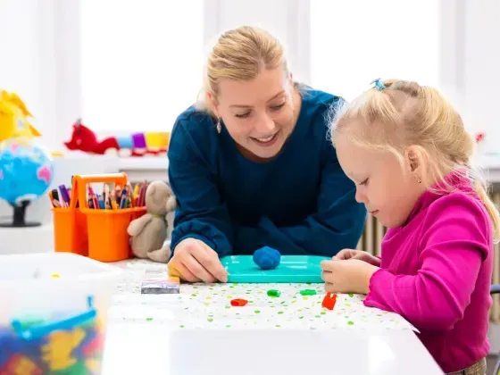 Woman helps a child play with colorful clay at a white table in a brightly lit room.