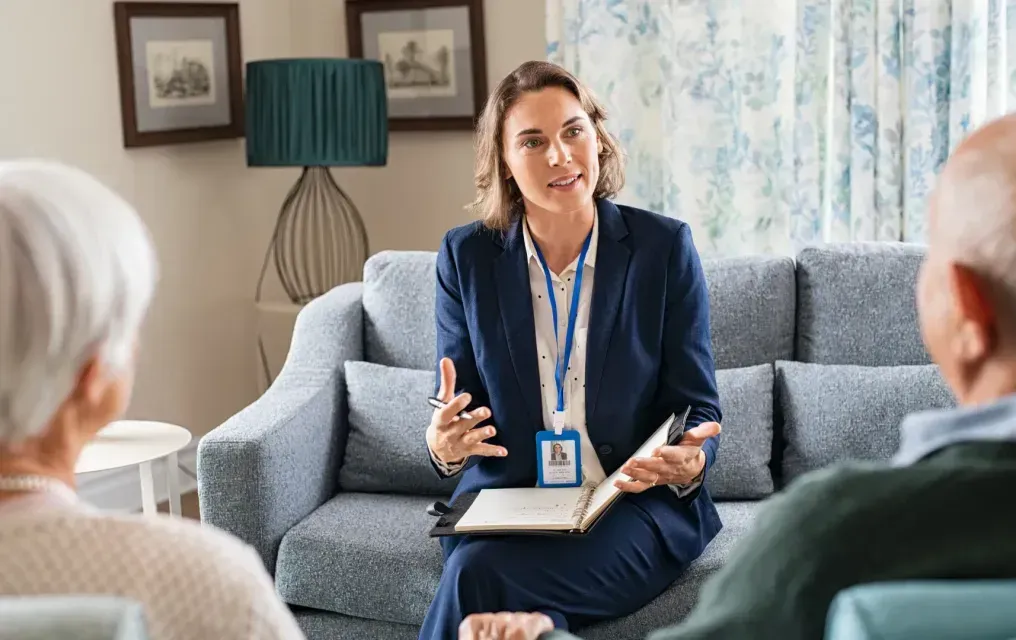 Woman in suit speaks to senior couple on a sofa, holding a notebook, indoors.