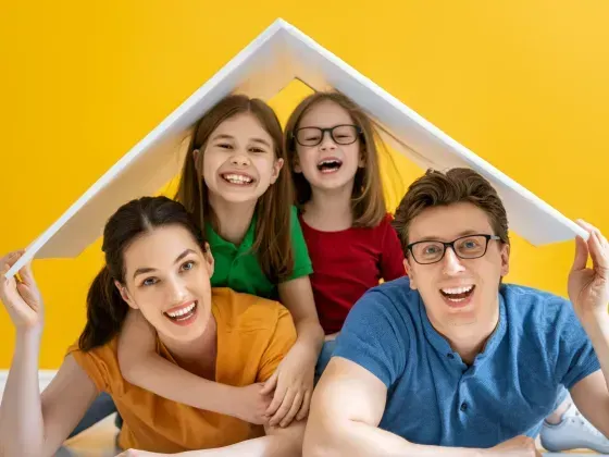 Family of four smiling, holding a white sheet to form a roof, against a yellow background.