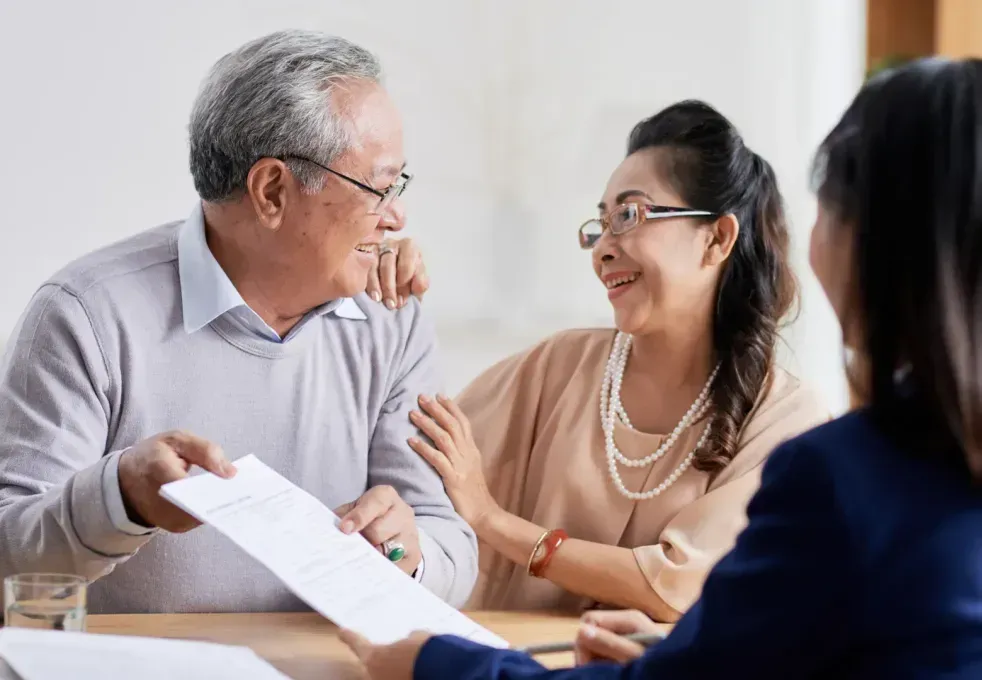 Older couple smiles at a person as they review paperwork at a table.