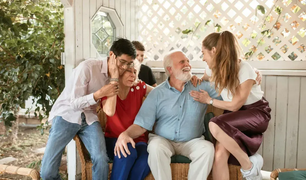 Family on a porch laughing and hugging; one man in blue shirt, woman in red top.