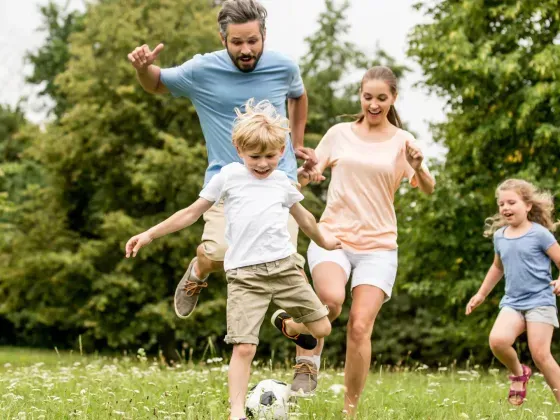Family playing soccer in a grassy park: parents jumping, kids running, ball in play, sunny day.