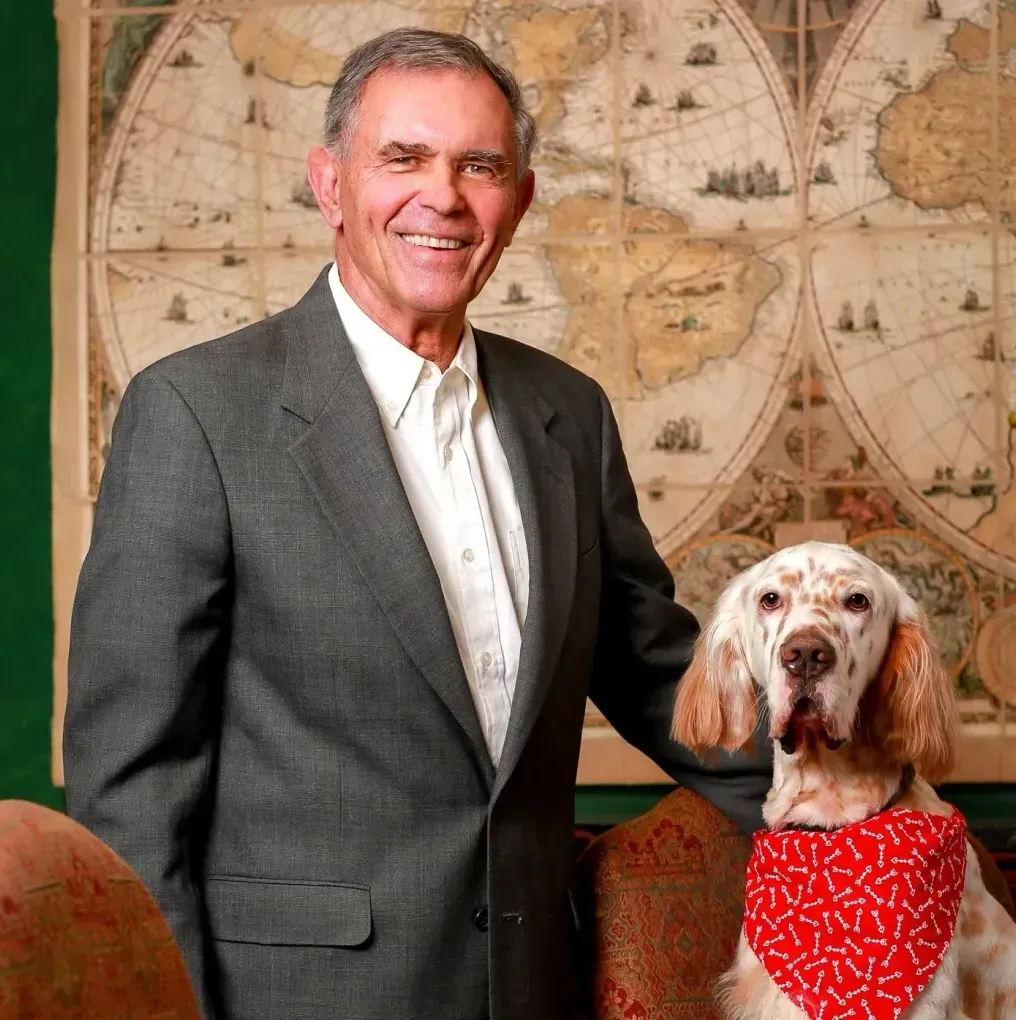 Man in suit smiles, standing with a dog wearing a bandana, in front of a world map.