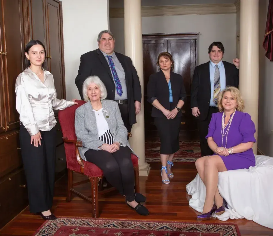 Group of people posing in an interior. Men and women wear business attire. Setting has pillars, wood cabinetry and rug.
