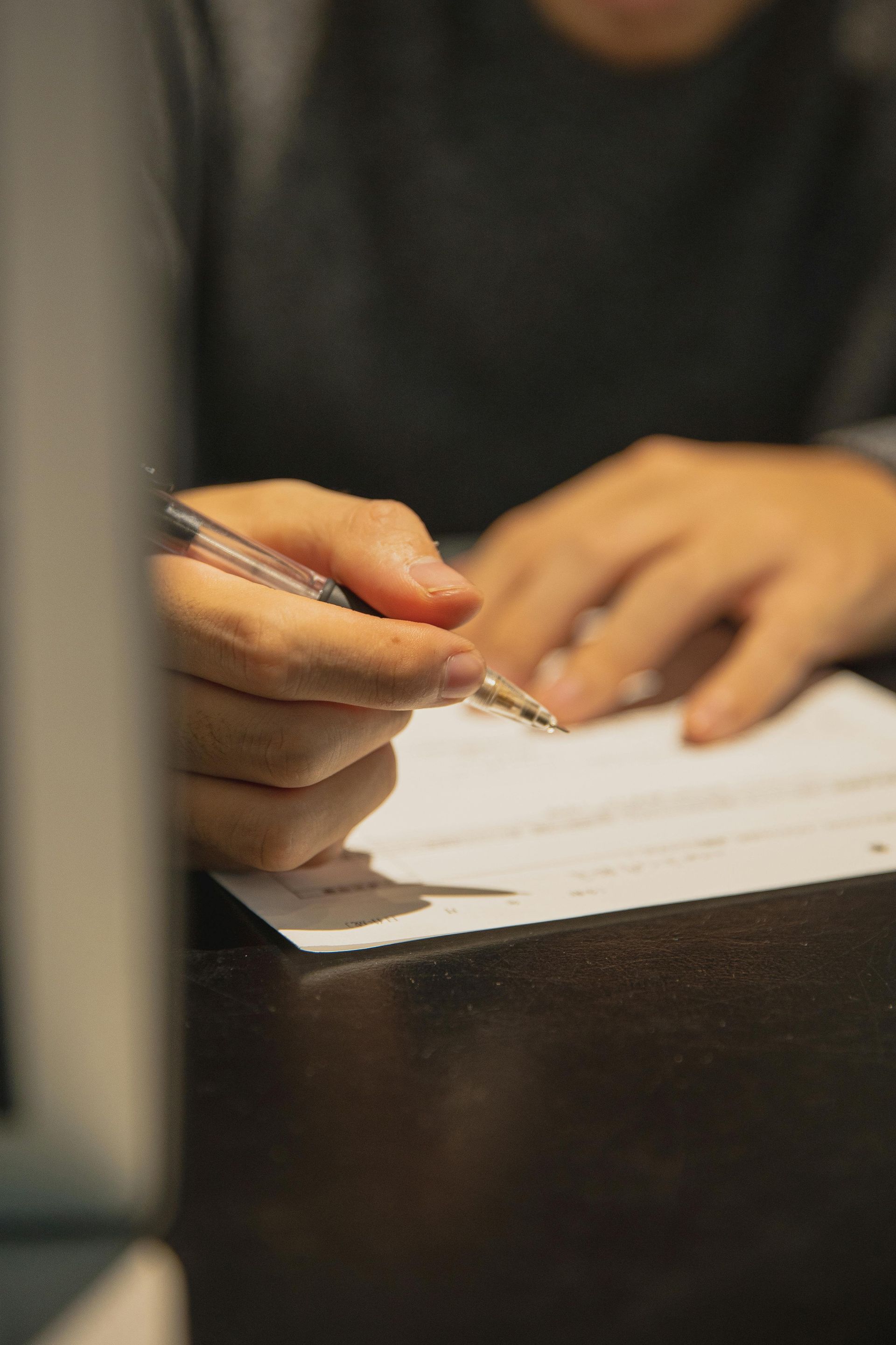 Person writing on a white paper with a clear pen at a dark desk.
