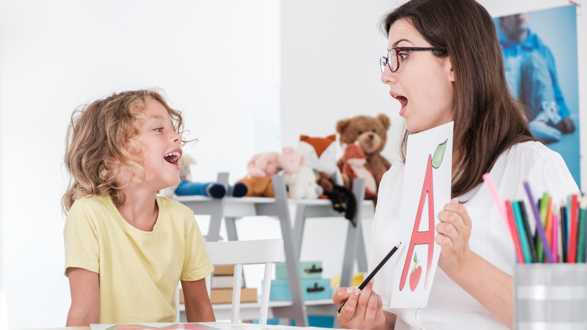 Therapist with glasses holds letter card, child imitates sound, in a room with toys and art supplies.