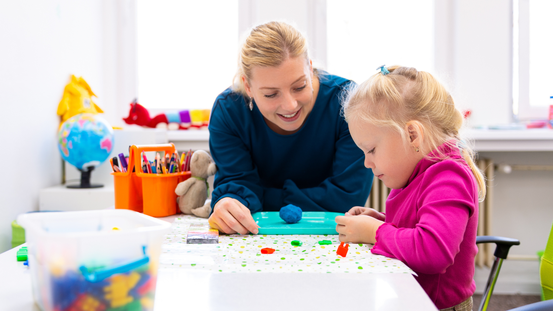 Woman helps a child play with colorful clay at a white table in a brightly lit room.