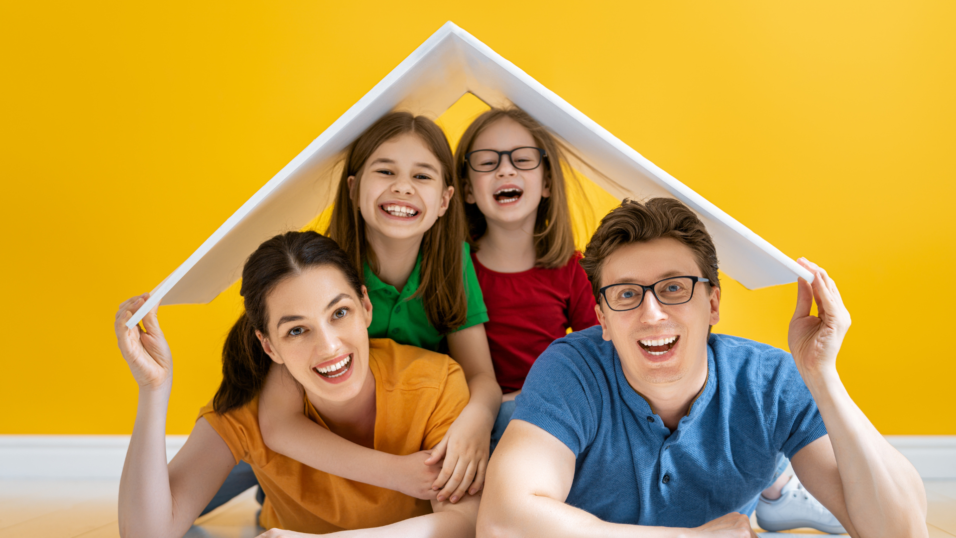 Family of four smiling, holding a white sheet to form a roof, against a yellow background.