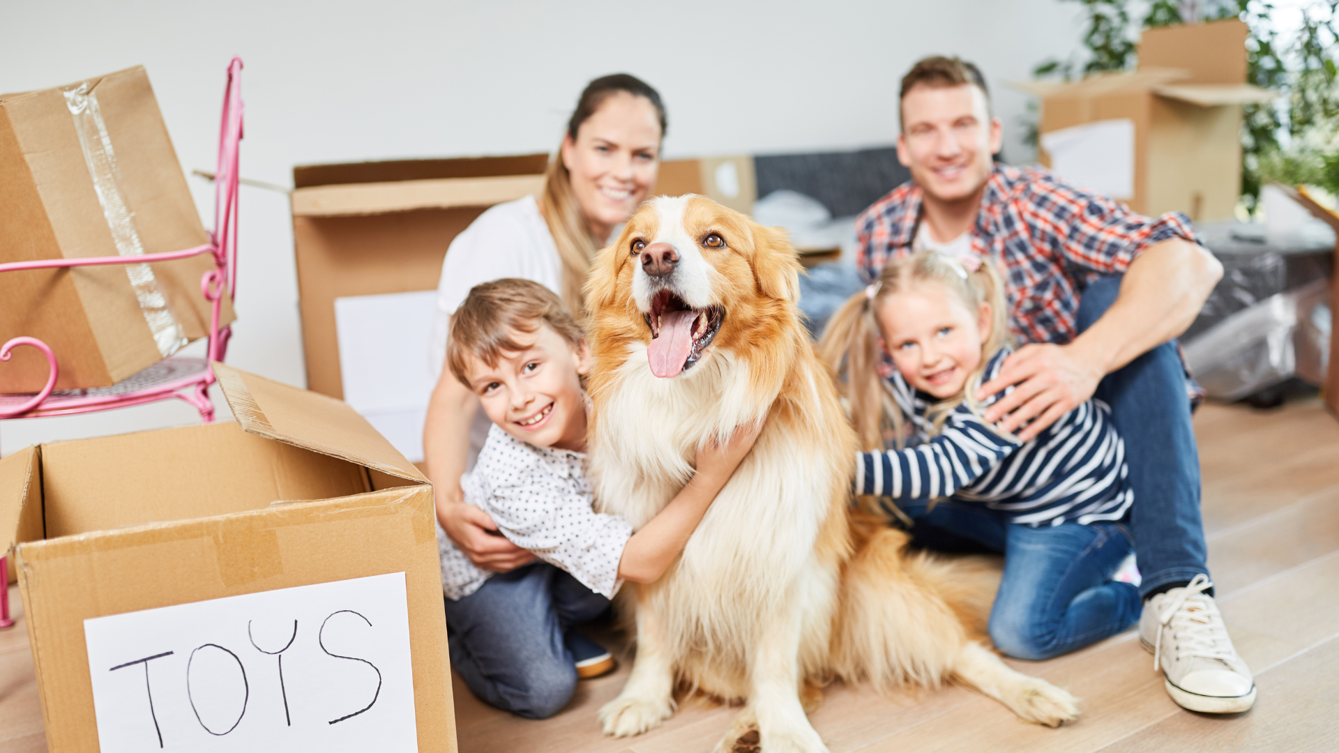 Family with two children and a dog surrounded by moving boxes, smiling.