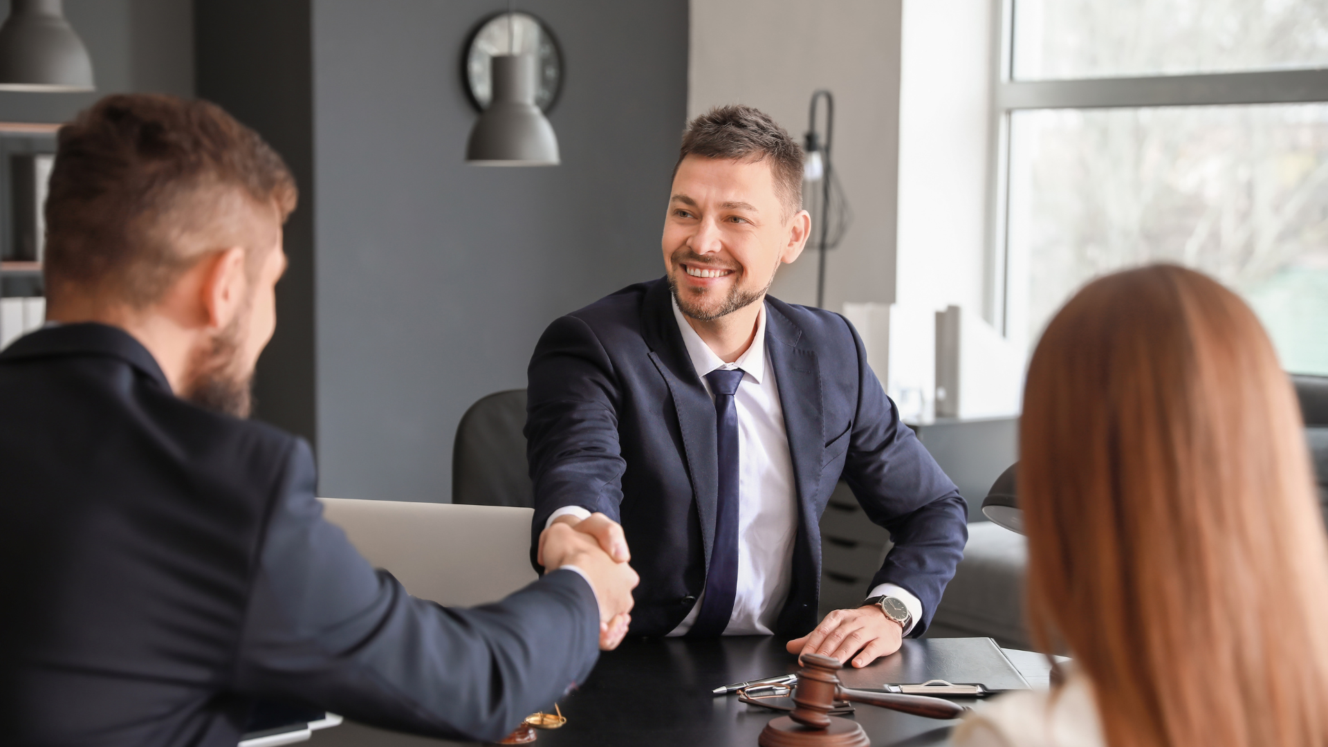 Two men in suits shaking hands at a table, smiling, with a woman in the background.