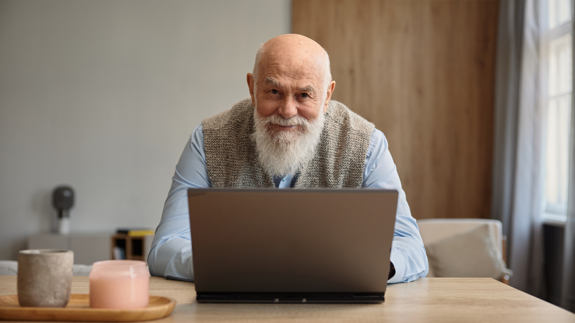 Older man with long white beard using a laptop at a table indoors, smiling.