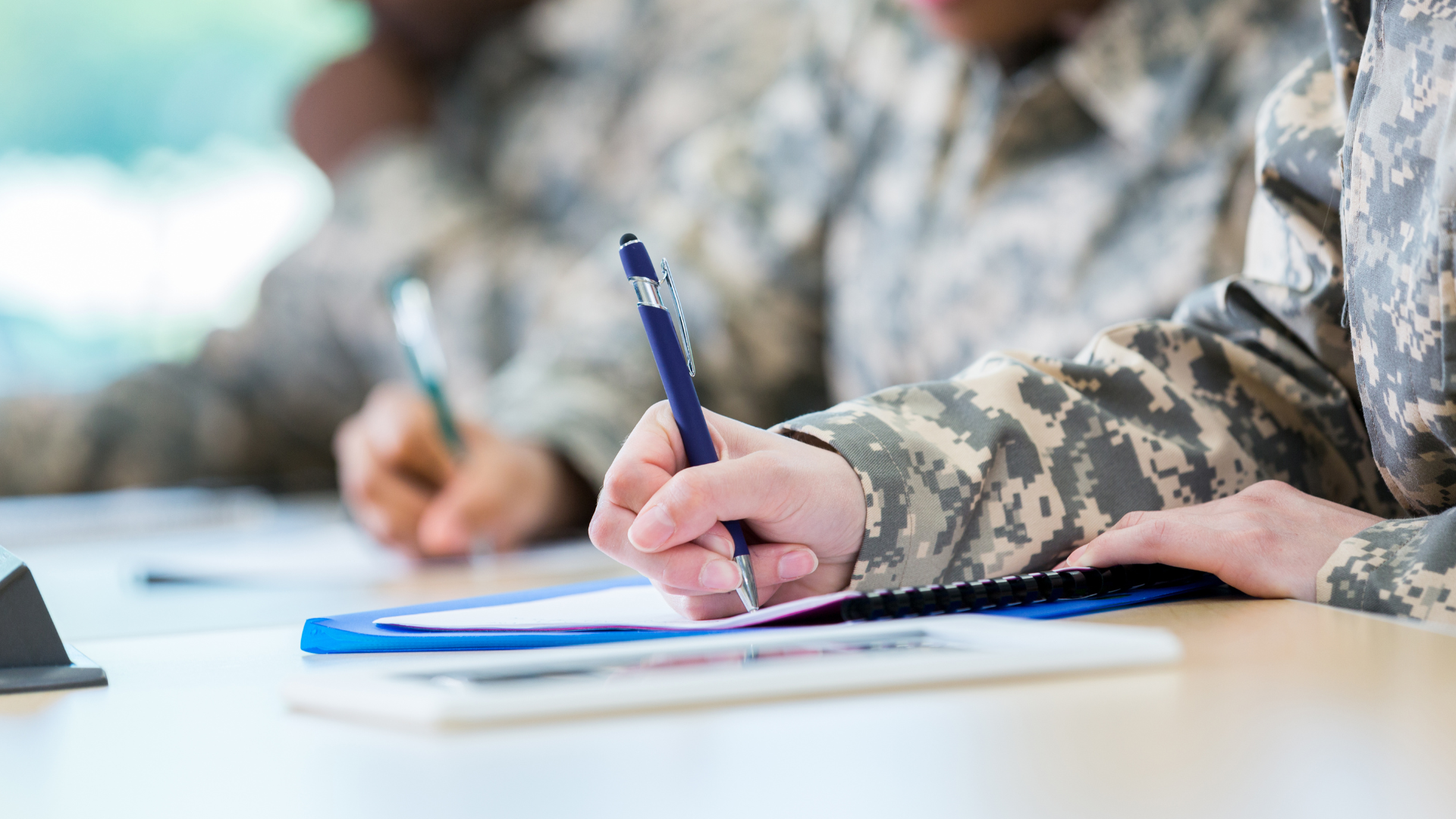 People in camouflage writing on paper, possibly in a meeting or training.