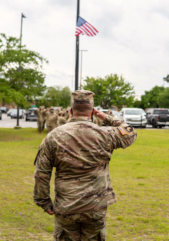 Soldier saluting American flag at a flagpole; other soldiers in background on a grassy field.